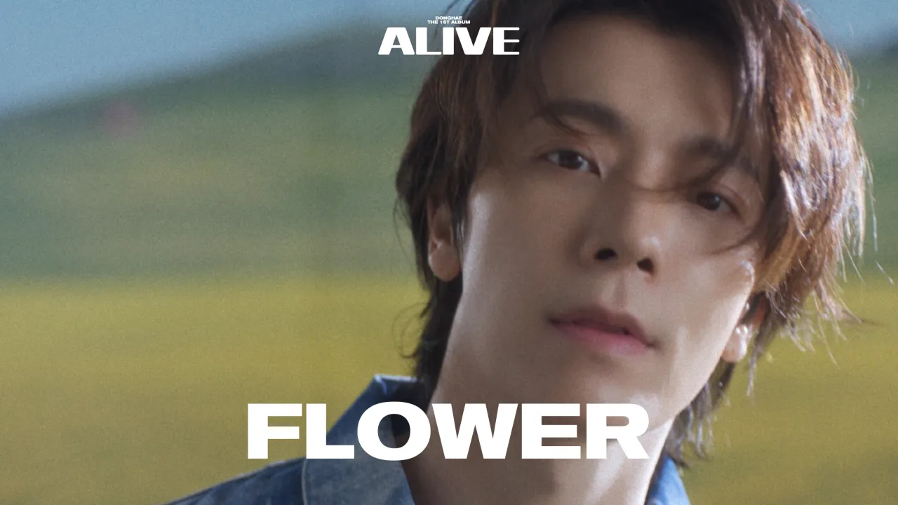 Still from the Flower music video — DONGHAE in a denim shirt before a sunlit yellow field, under the album's ALIVE wordmark with the FLOWER title across the frame